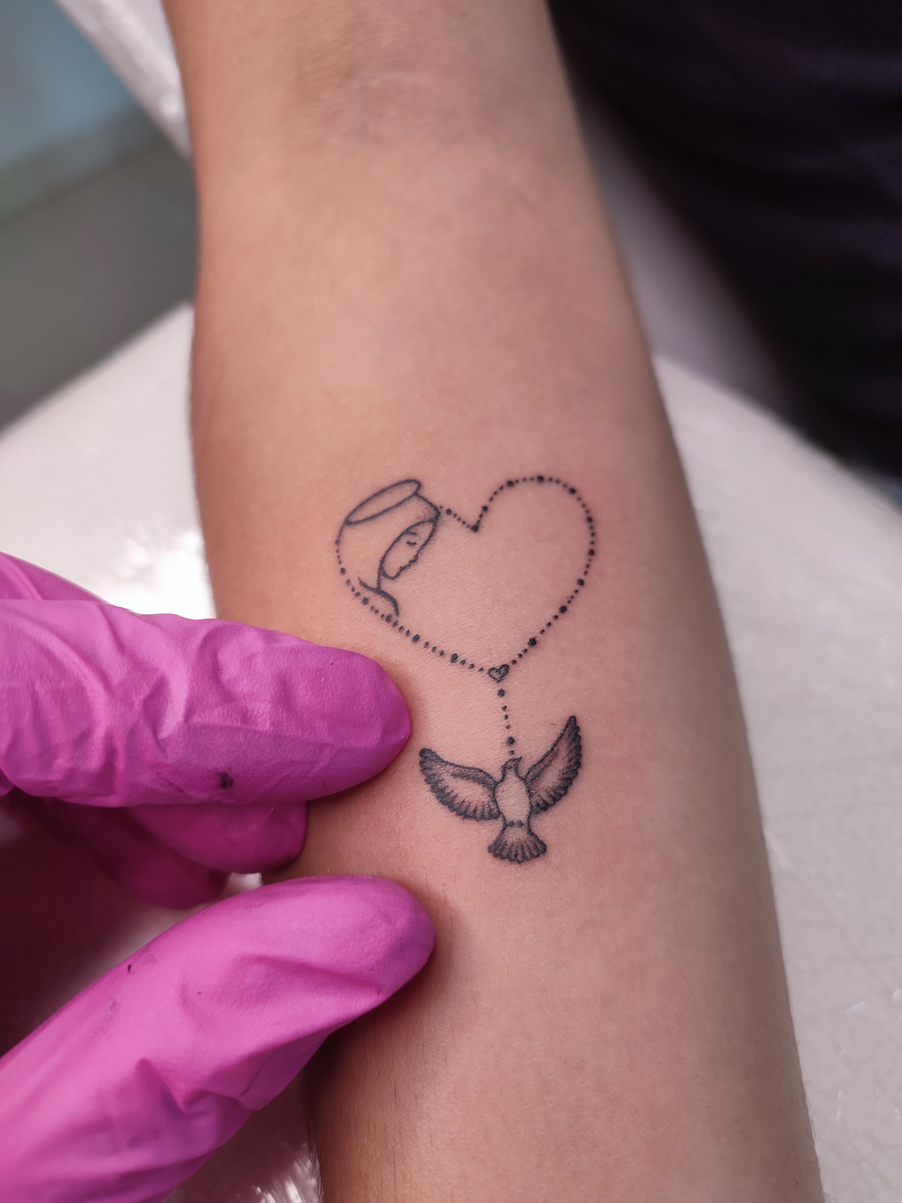 Tatuagem minimalista da nossa senhora dentro de um coraçao, com a ponta do coraçao como um terço que no final tem uma pomba da paz, representando o espirito santo